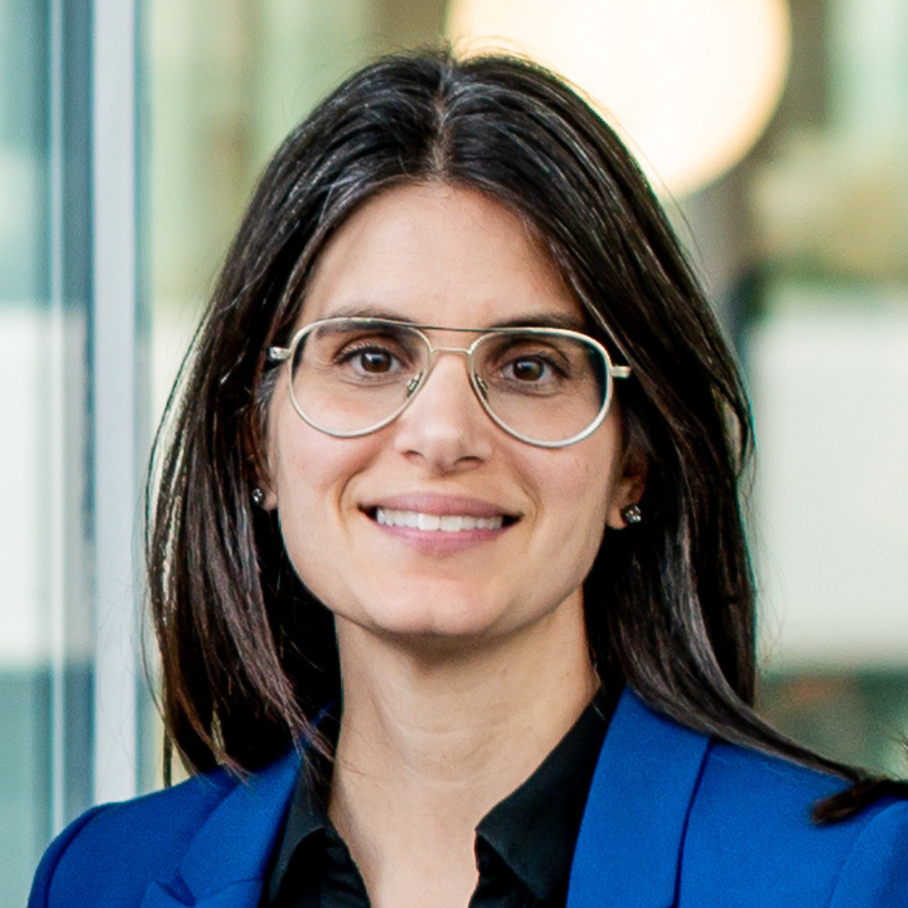 A professional individual with dark hair and glasses, dressed in a blue blazer, smiling confidently in a modern environment.