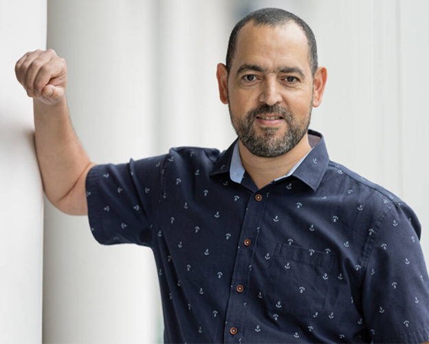 A confident individual with a friendly smile leans against a wall, dressed in a casual, patterned shirt.