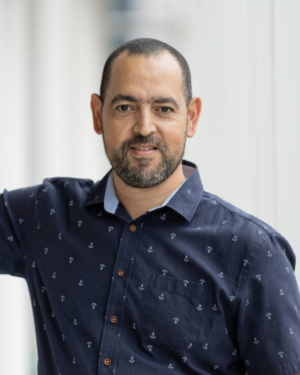 Un homme souriant en chemise bleue à motifs, posant avec confiance dans un environnement moderne et lumineux.