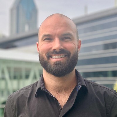 A smiling individual with a beard, wearing a black shirt, stands in an urban environment featuring modern architecture.