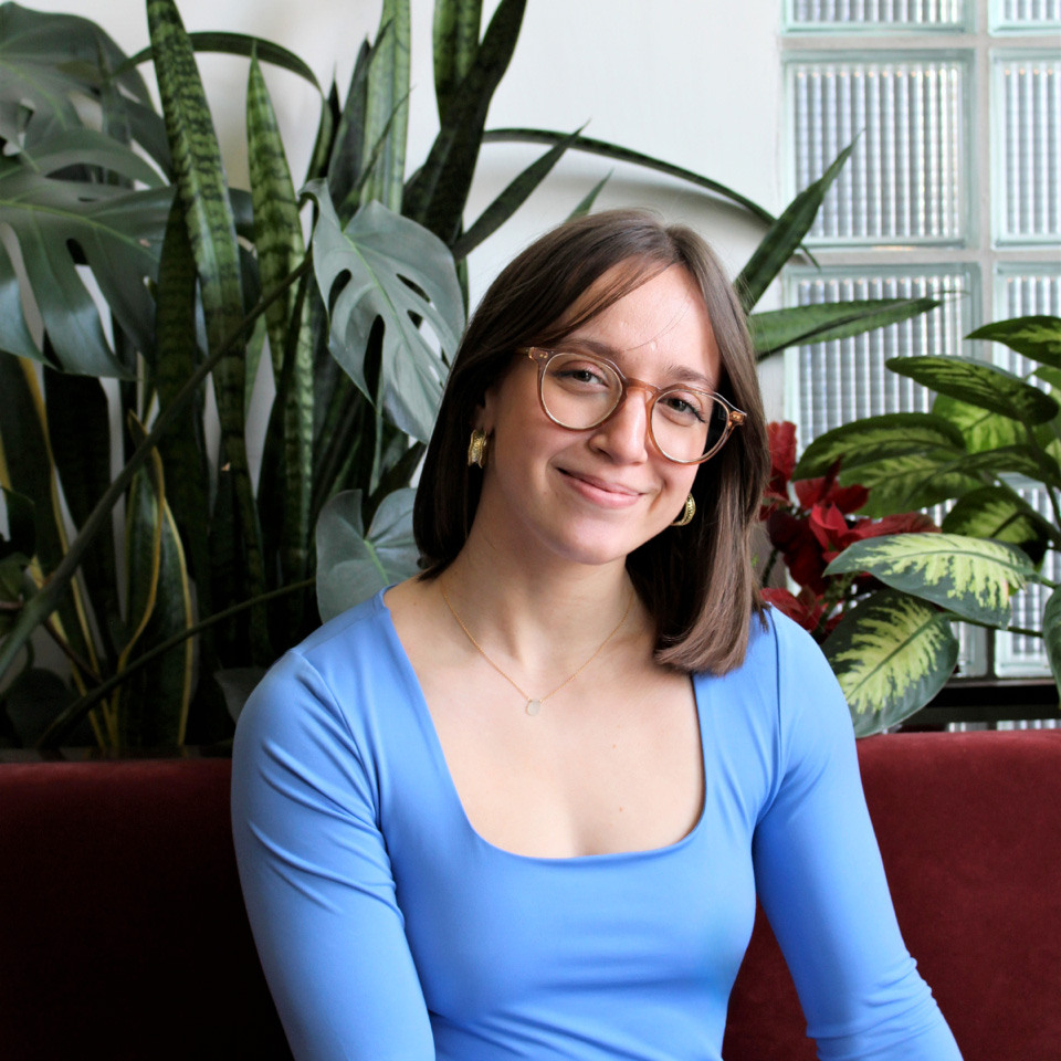 Étudiante souriante vêtue d'un top bleu, entourée de plantes, évoquant un environnement accueillant et inspirant.