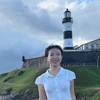 Une jeune femme souriante se tient devant un phare emblématique, avec une colline verdoyante et un ciel nuageux en arrière-plan.