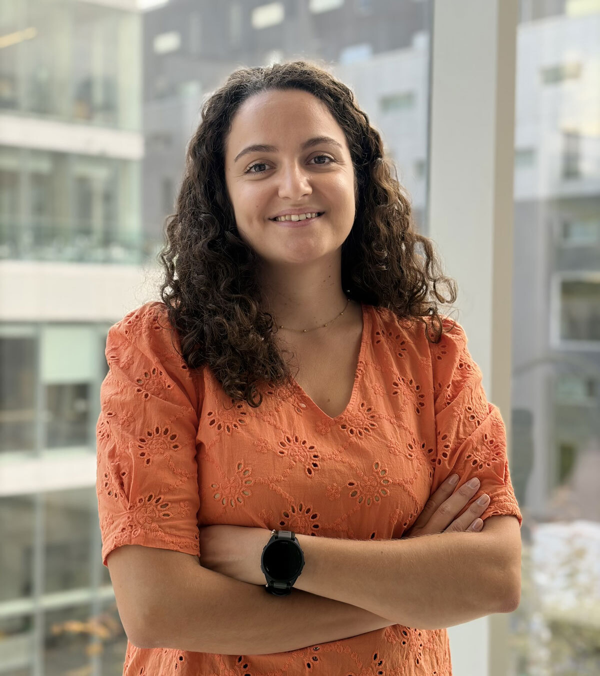 Portrait d'une jeune femme souriante, portant un haut orange, avec les bras croisés, se tenant devant une grande fenêtre au sein d'un campus moderne.