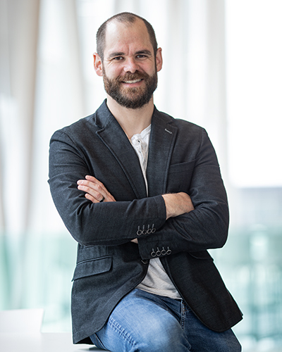A professional smiling individual with a beard, wearing a blazer and casual attire, posing confidently in a modern, bright environment.