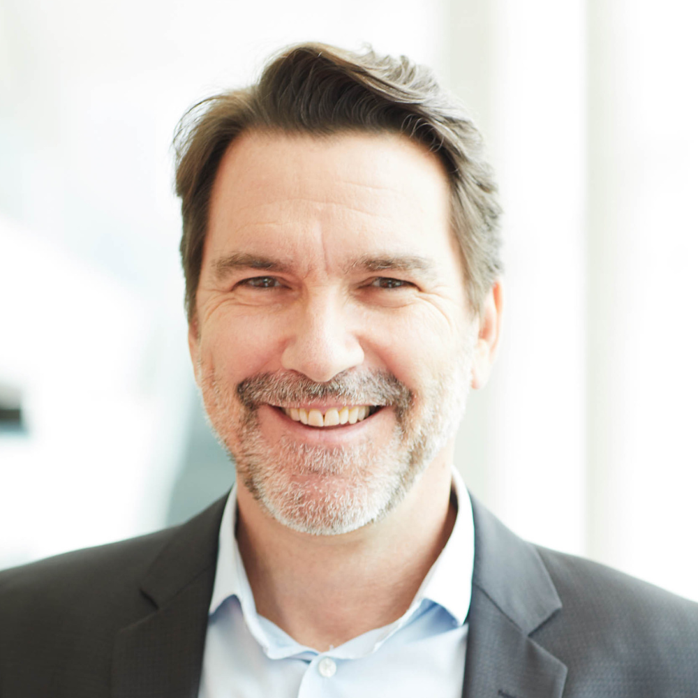 A professional man with salt-and-pepper hair and a warm smile, wearing a brown sweater, embodies a welcoming academic environment.