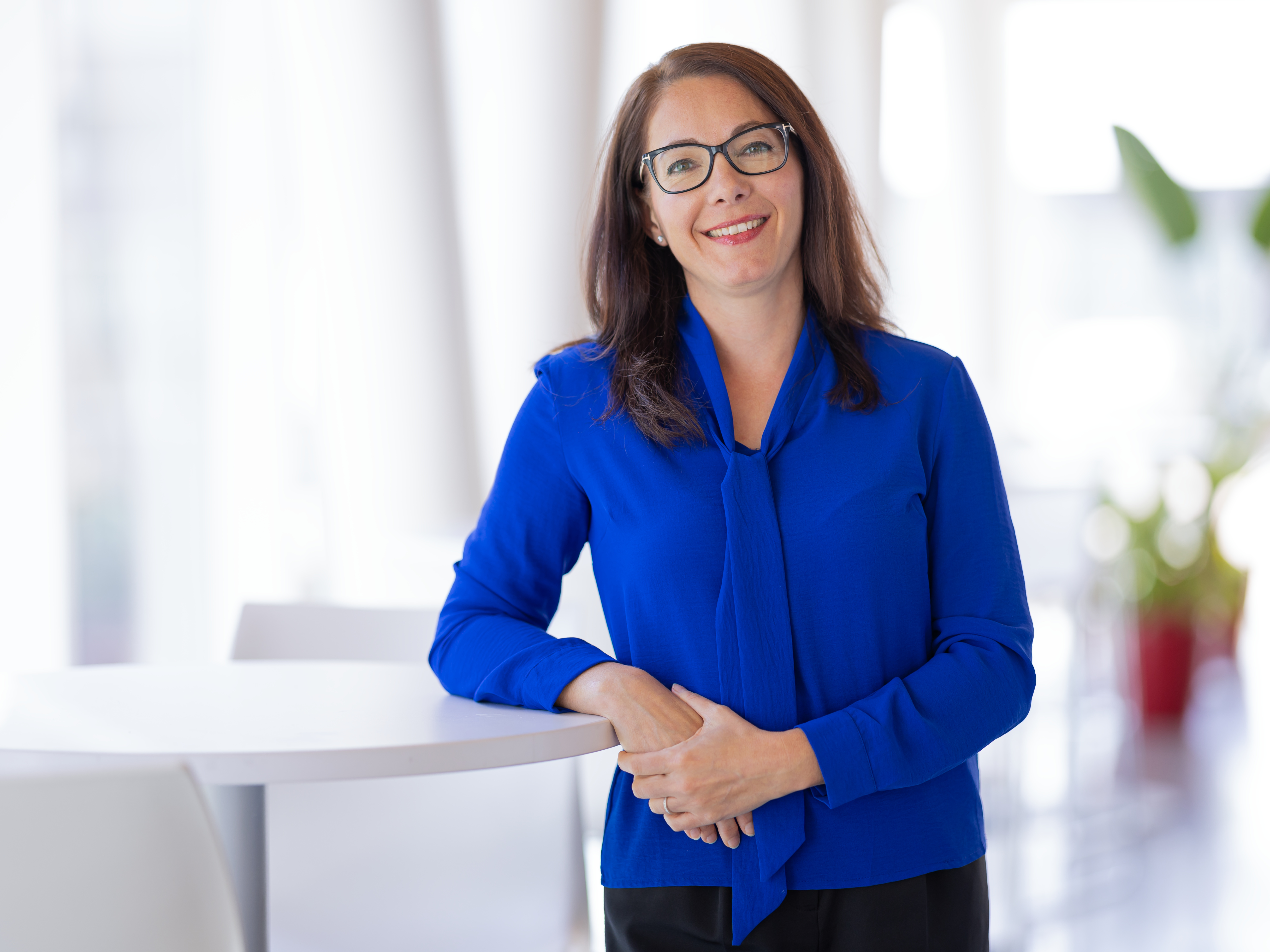 Une professionnelle souriante en chemisier bleu se tient derrière une table, prête à accueillir et à partager ses connaissances.