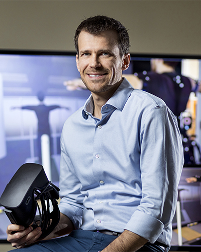 Un chercheur souriant, tenant un casque de réalité virtuelle, se tient devant un écran montrant des applications technologiques innovantes.