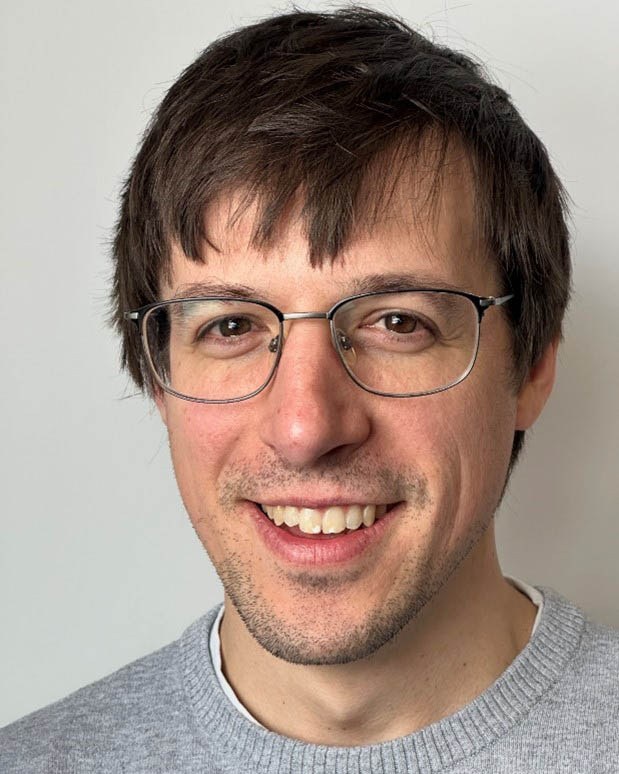 Portrait d'un homme souriant, présentant des lunettes et portant un pull gris, affichant une expression amicale et engageante.