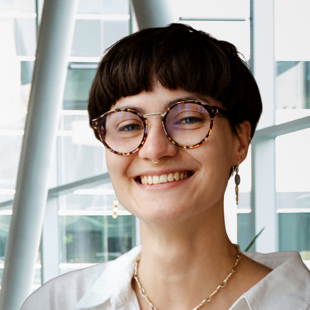 A smiling student with short hair and glasses stands in a modern, well-lit space, showcasing a positive and friendly demeanor.