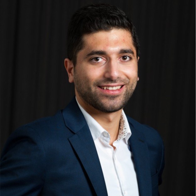 Un jeune homme souriant en costume, portant une chemise blanche, se tient devant un fond noir. Ambiance professionnelle et amicale.