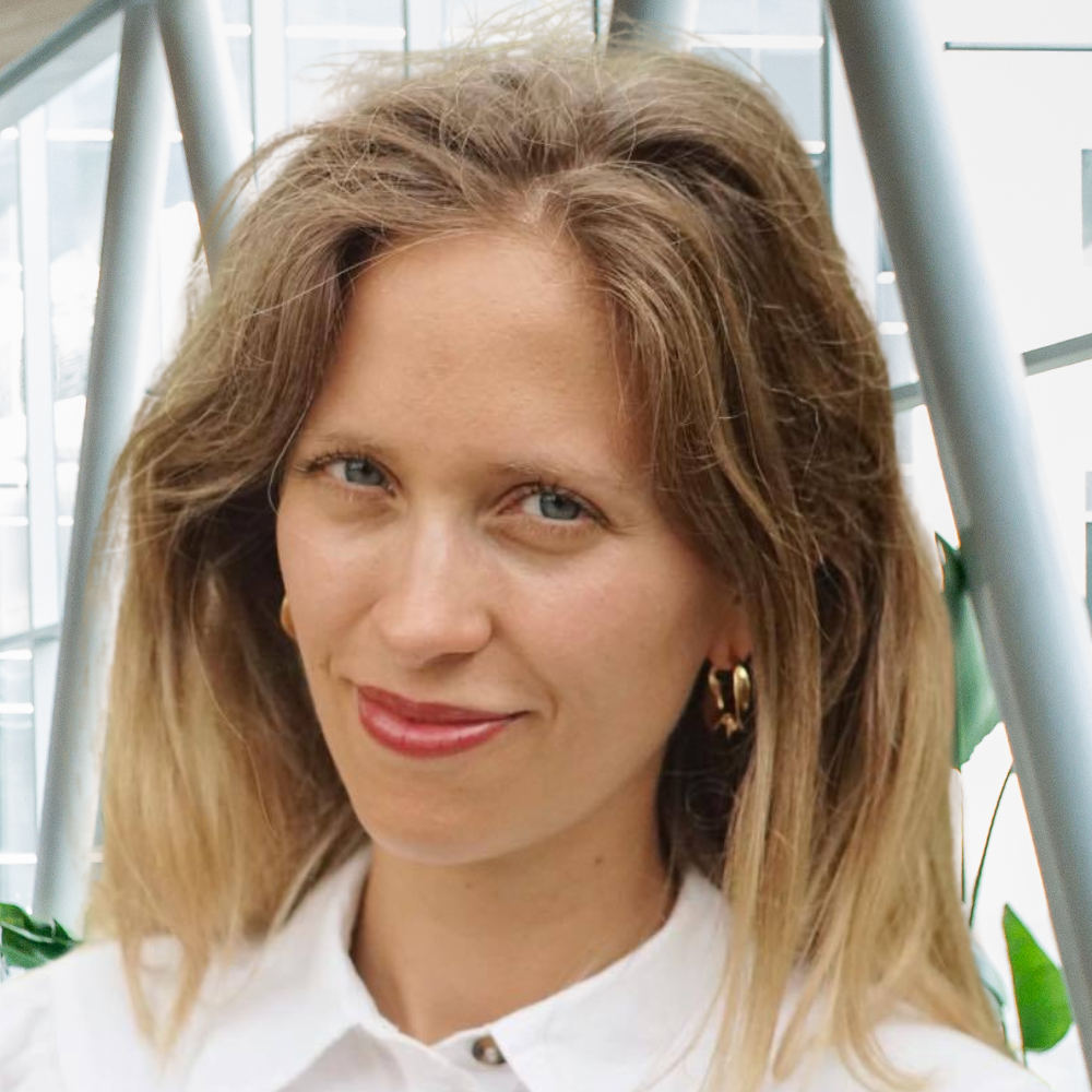 A woman with a friendly smile is wearing a white shirt and gold earrings, surrounded by greenery and modern architecture.