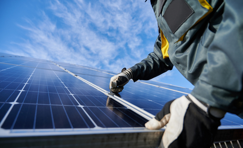 Les bras d'un technicien portant des gants de travail en train d'installer un système photovoltaïque de panneaux solaires autonome sous un beau ciel bleu avec des nuages.