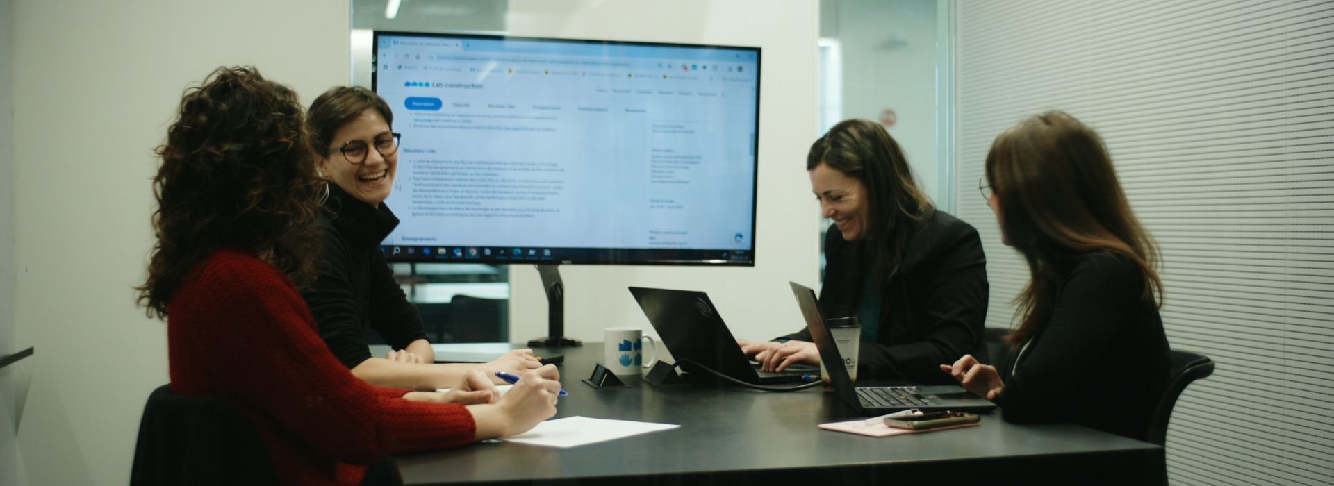 Quatre personnes échangent autour d'une table, collaborant sur un projet avec des ordinateurs et un écran affichant des informations.