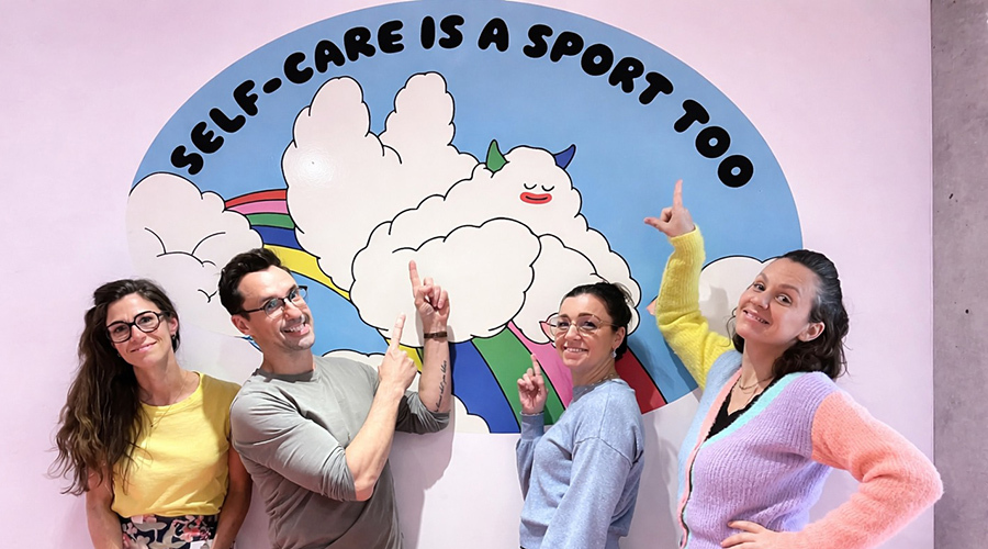 Quatre personnes amusées signalent une fresque colorée au message inspirant sur le bien-être et l'importance de prendre soin de soi.