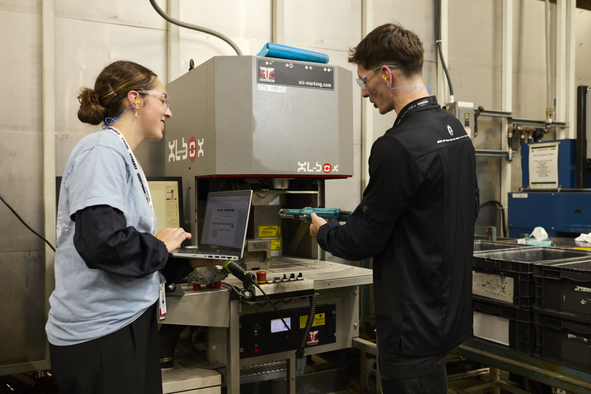 Deux étudiants collaborent dans un laboratoire technique, utilisant un ordinateur et un équipement de marquage. Sécurité prioritaire.