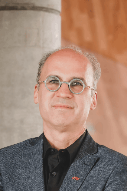 Portrait d’un homme debout devant un arrière-plan architectural, vêtu d’une veste foncée et de lunettes rondes, regard face caméra.