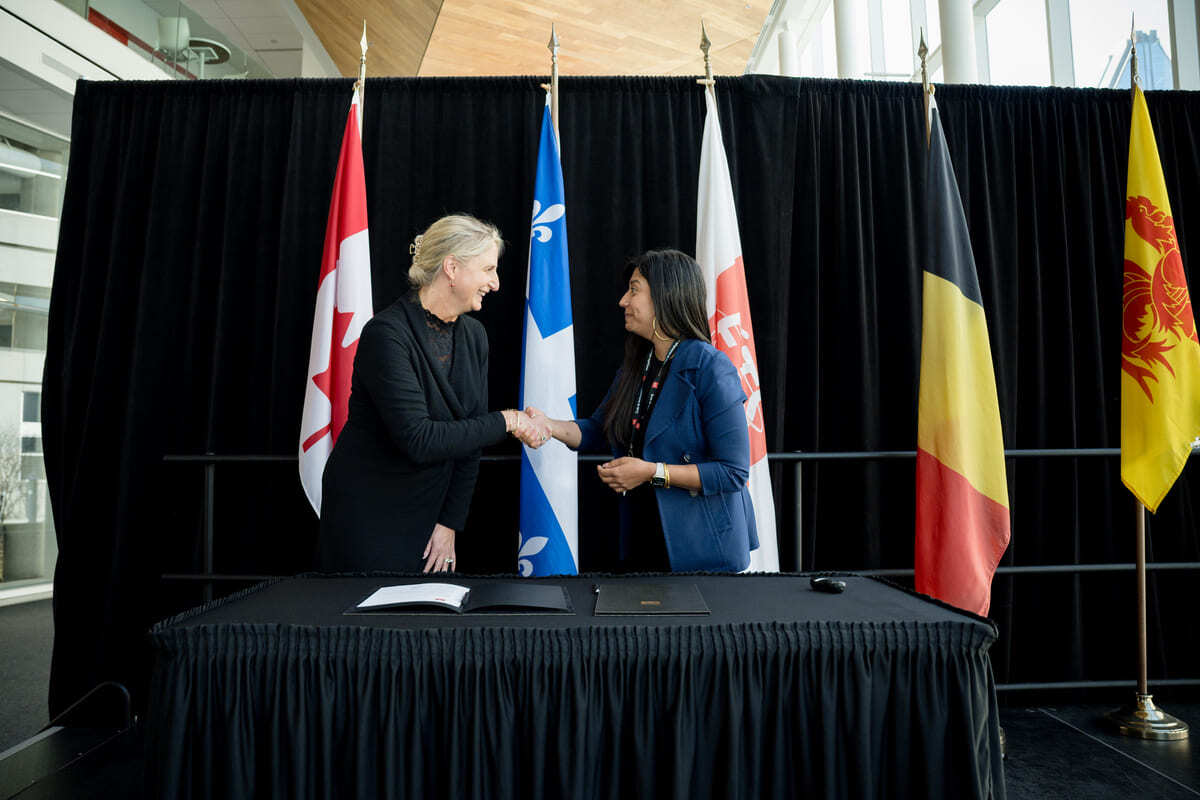 Deux femmes se serrent la main lors d'un événement officiel, entourées de drapeaux du Canada et du Québec.