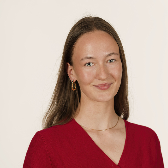 Une jeune femme souriante portant un haut rouge, avec des boucles d'oreilles dorées et des cheveux longs, dégage une attitude confiante.