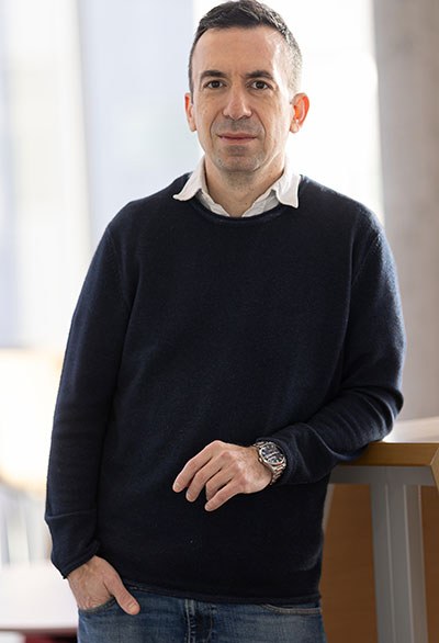 A confident individual stands by a modern workspace, wearing a dark sweater and casually positioned, conveying a professional demeanor.