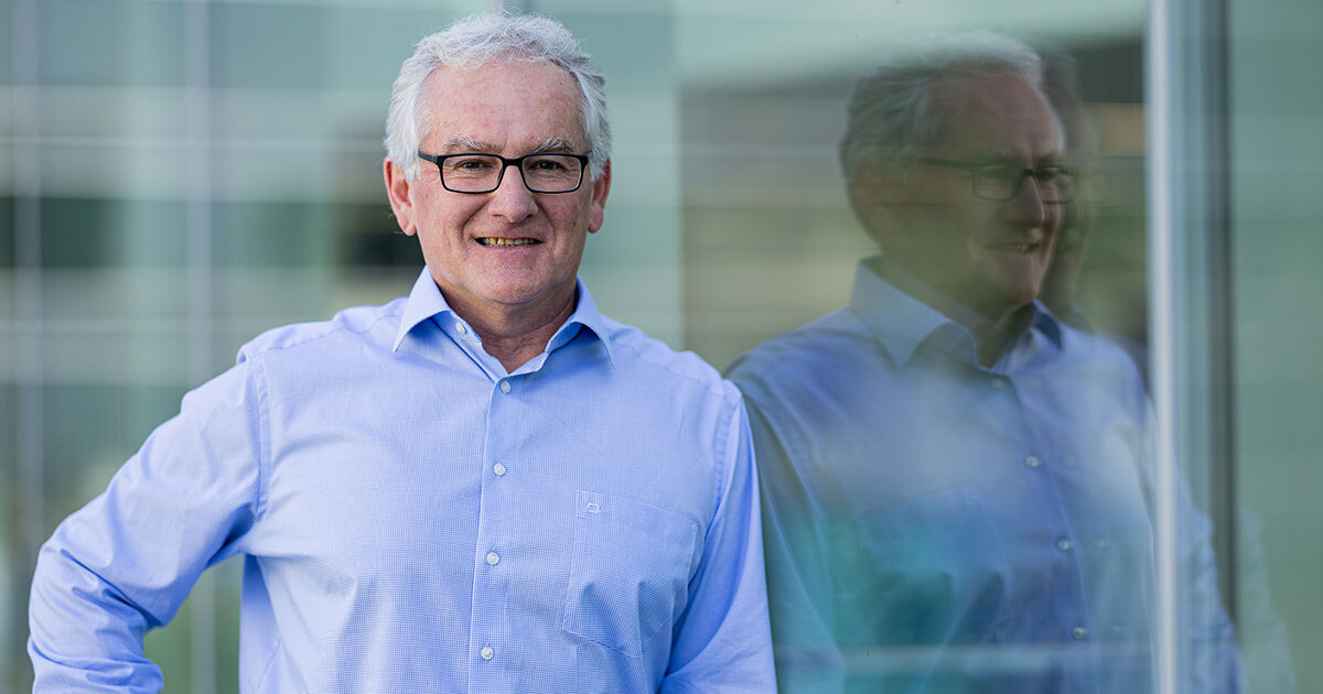 Un homme souriant porte une chemise bleue, adossé à une surface vitrée, reflétant un environnement moderne et lumineux.