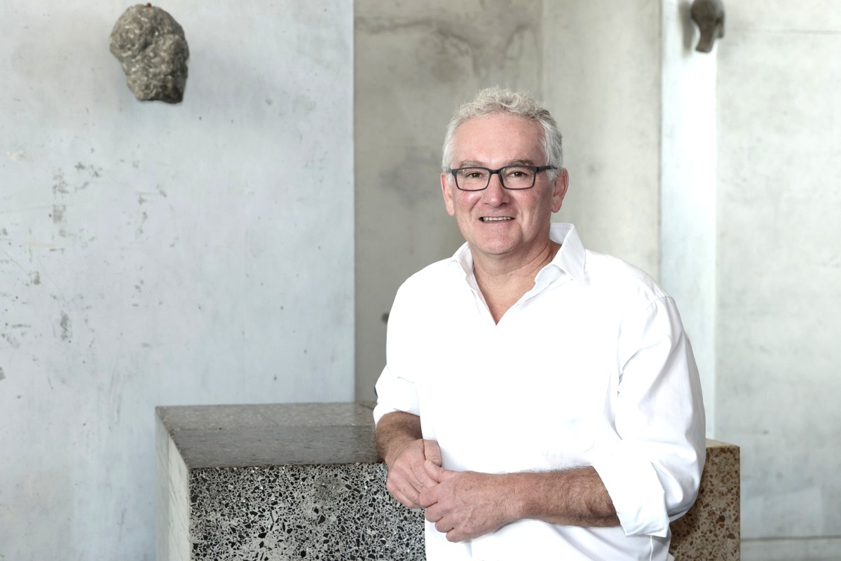 Un professeur souriant, vêtu d'une chemise blanche, se tient dans un espace contemporain avec des éléments en béton.