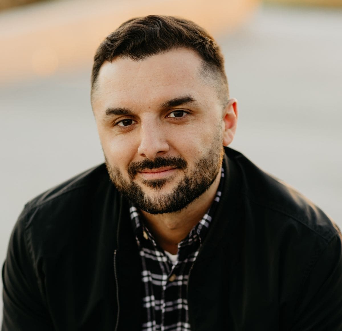 Un homme souriant, vêtu d'une chemise à carreaux et d'une veste noire, dégage une attitude attentive et sympathique.