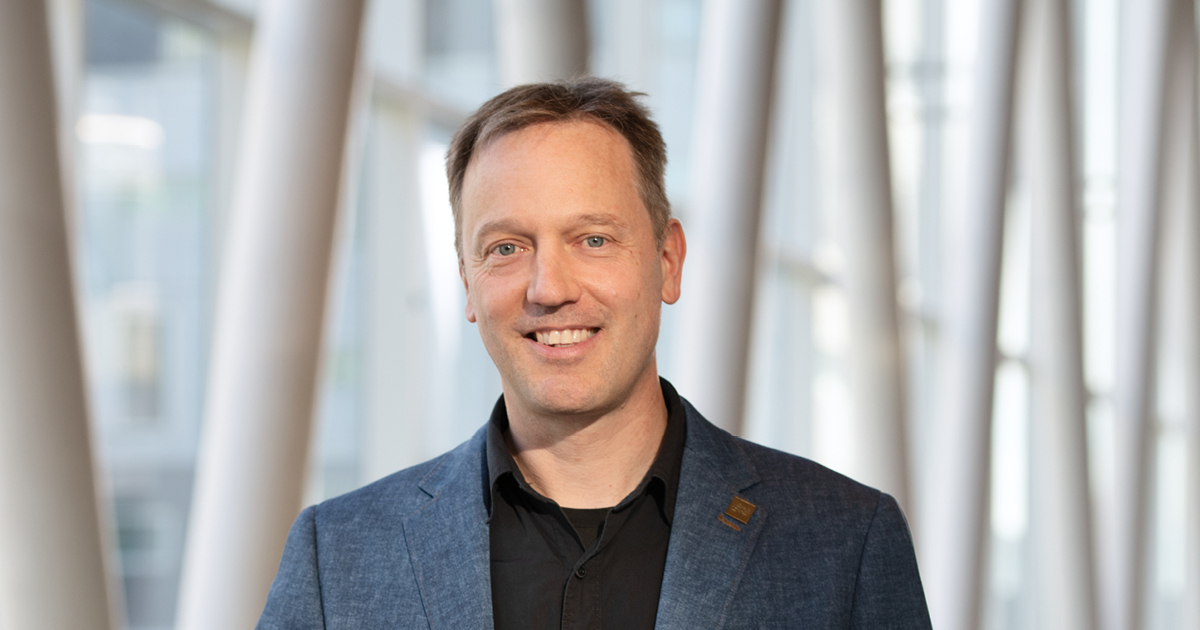Professeur en technologie, souriant, portant un blazer, devant un fond architectural moderne, reflétant l'innovation et l'expertise.