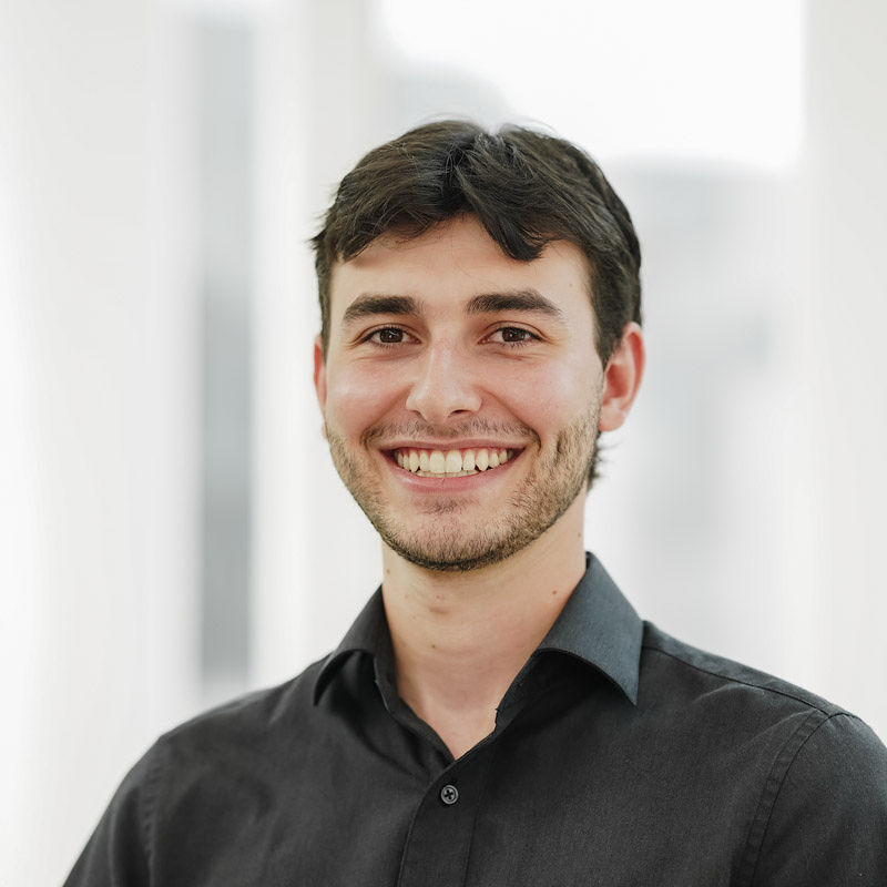 Un jeune homme souriant en chemise noire, se tenant debout dans un environnement lumineux et moderne.