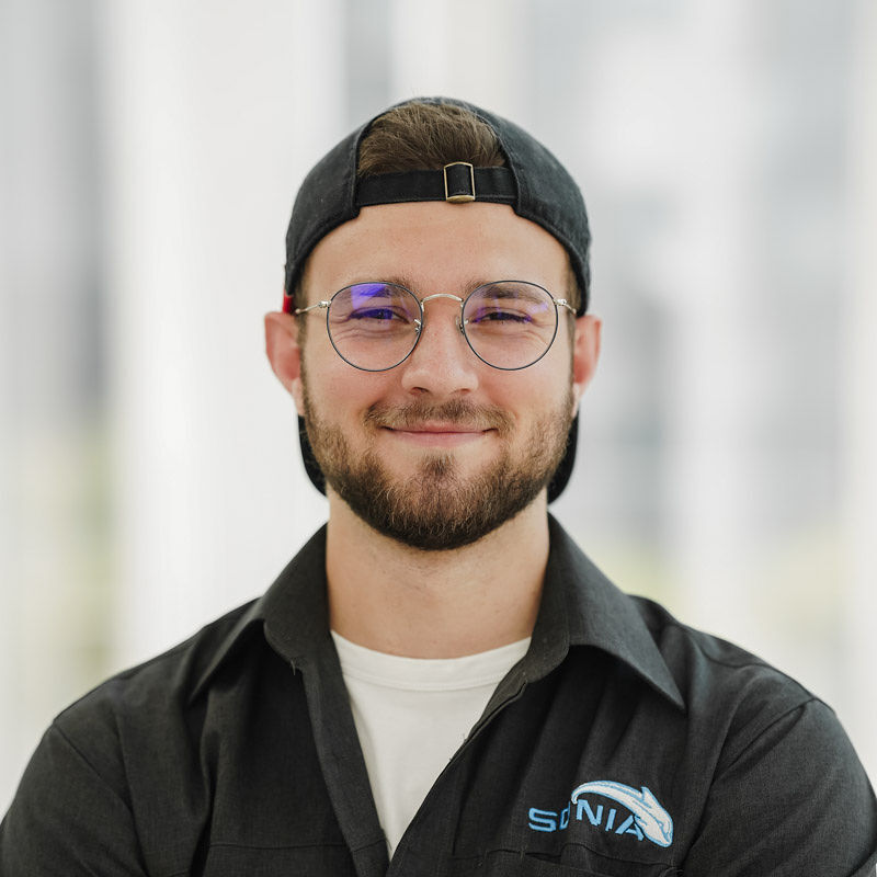 Un étudiant souriant portant des lunettes, une casquette, et un uniforme professionnel, dans un environnement moderne et lumineux.
