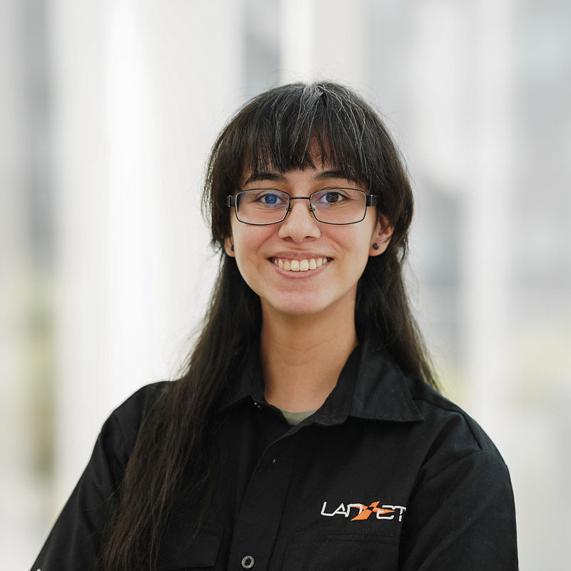 Étudiante souriante en blouse noire, passionnée par la technologie, prête à relever des défis académiques et professionnels.