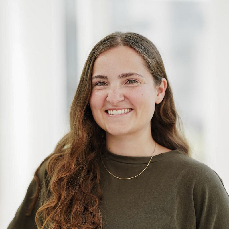 Sourire franc et chaleureux, des cheveux longs et ondulés, exprimant confiance et positivité dans un environnement académique moderne.