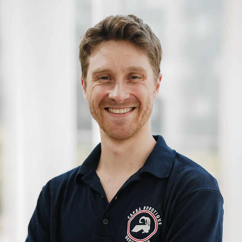 Un homme souriant porte un polo bleu avec un logo, se tenant dans un environnement lumineux et moderne.