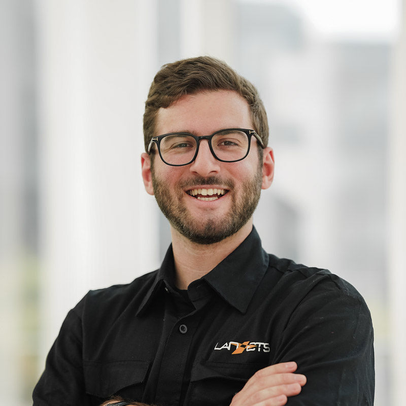 Un étudiant souriant, vêtu d'une chemise noire, affiche confiance et enthousiasme dans un environnement moderne et lumineux.