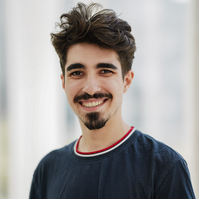 Un étudiant souriant, aux cheveux bouclés et à la barbe soignée, dégage une atmosphère positive et accueillante.