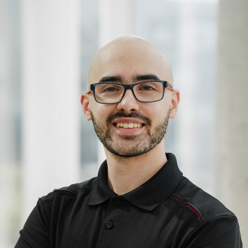 Étudiant souriant, portant une chemise noire, se tient en confiance dans un environnement moderne et lumineux.