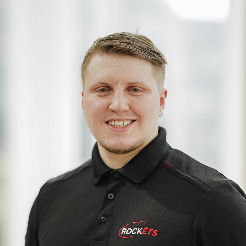Un étudiant souriant porte un polo noir avec le logo "ROCKETS", exprimant enthousiasme et professionnalisme dans un cadre moderne.