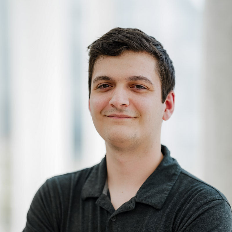 Un étudiant souriant, debout avec les bras croisés, dégage une atmosphère confiante et amicale dans un environnement lumineux.