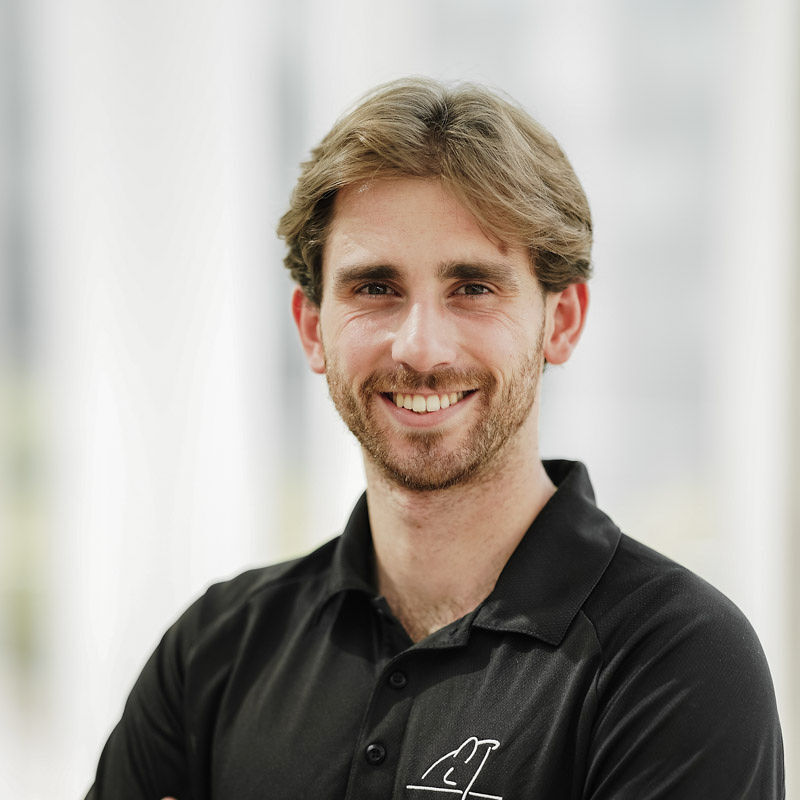 Un jeune homme souriant porte un polo noir et pose avec assurance, montrant un esprit accueillant et professionnel.
