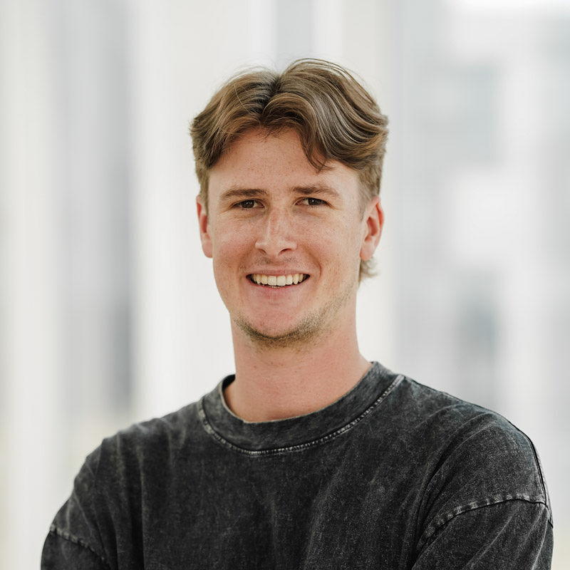Jeune homme souriant, portant un sweat noir, se tenant dans un environnement lumineux, évoquant convivialité et engagement.