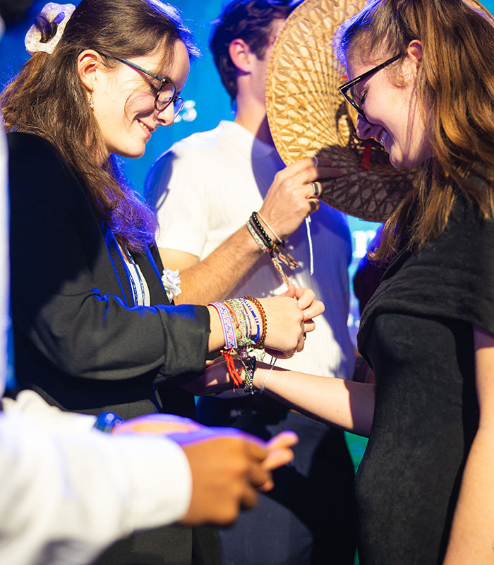 Deux jeunes femmes échangent des bracelets, entourées par un groupe, dans une ambiance festive et conviviale.