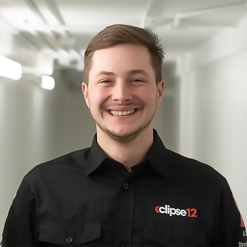 Un étudiant souriant porte une chemise noire avec le logo "eclipse12", dans un environnement moderne et éclairé.