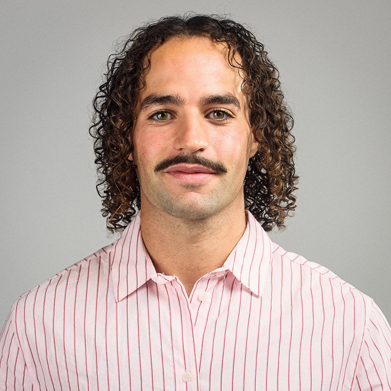 Un homme souriant avec des cheveux bouclés et une moustache, portant une chemise à rayures blanches et roses, sur fond neutre.