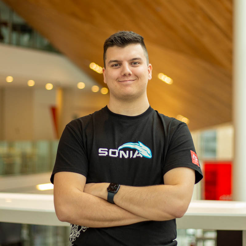 Étudiant confiant en technologie, il porte un t-shirt avec le logo SONIA, dans un environnement moderne et lumineux.