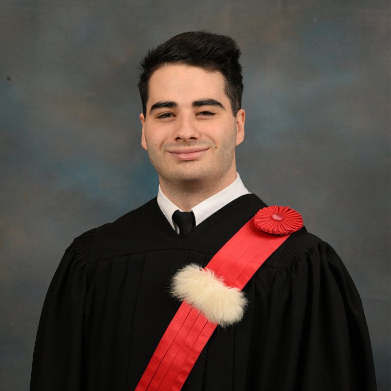Étudiant souriant en habit de remise de diplôme, avec un insigne rouge et blanc, marquant une étape importante de sa carrière académique.