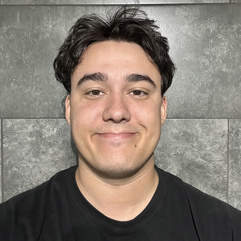Étudiant souriant, portant un t-shirt noir, devant un mur en carreaux gris. Énergie positive et attitude amicale.