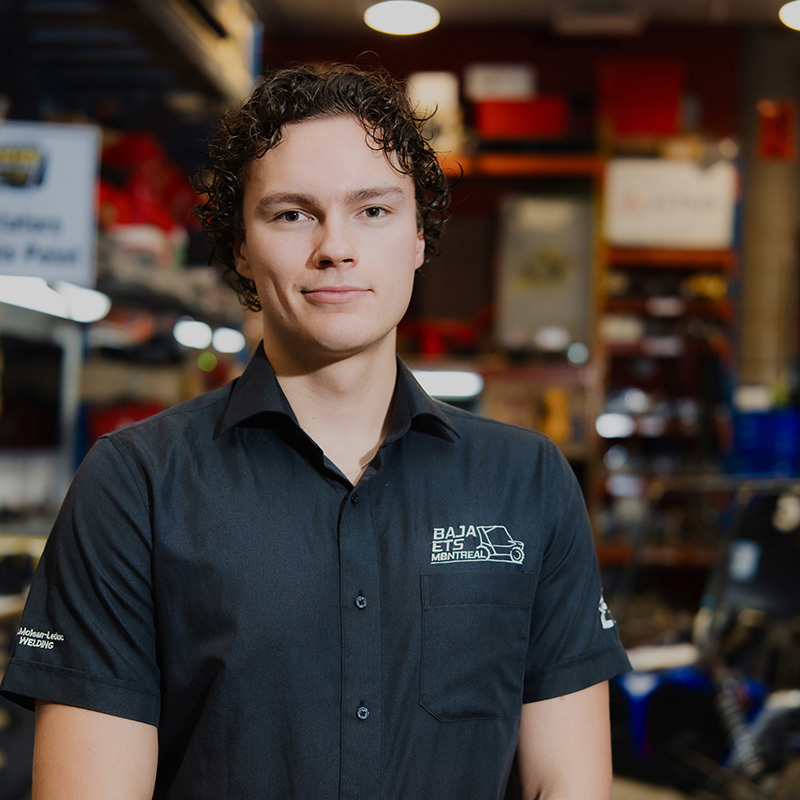 Un étudiant en technologie, souriant, porte une chemise noire dans un environnement d'atelier, entouré d'équipements techniques.