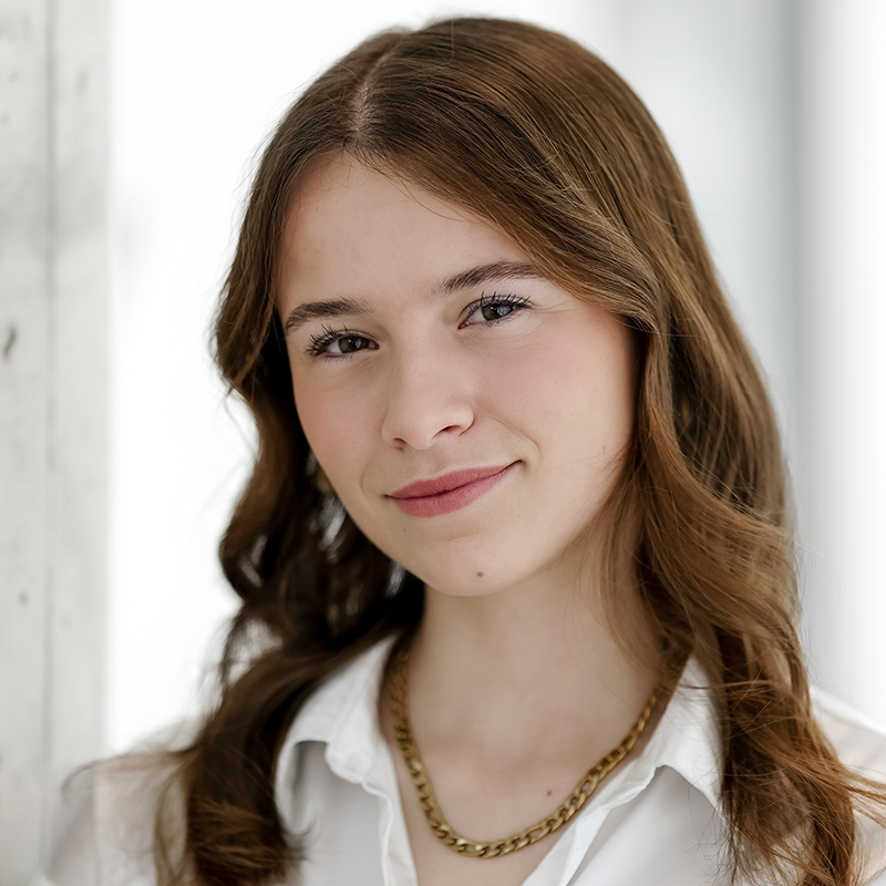 Jeune femme souriante avec des cheveux ondulés, portant une chemise blanche et un collier doré, dans un environnement lumineux.