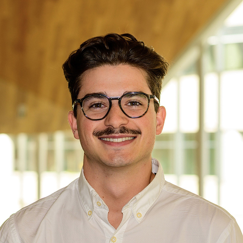 Jeune homme souriant, portant des lunettes, avec une chemise blanche dans un environnement moderne et lumineux. Ambiance amicale et accueillante.