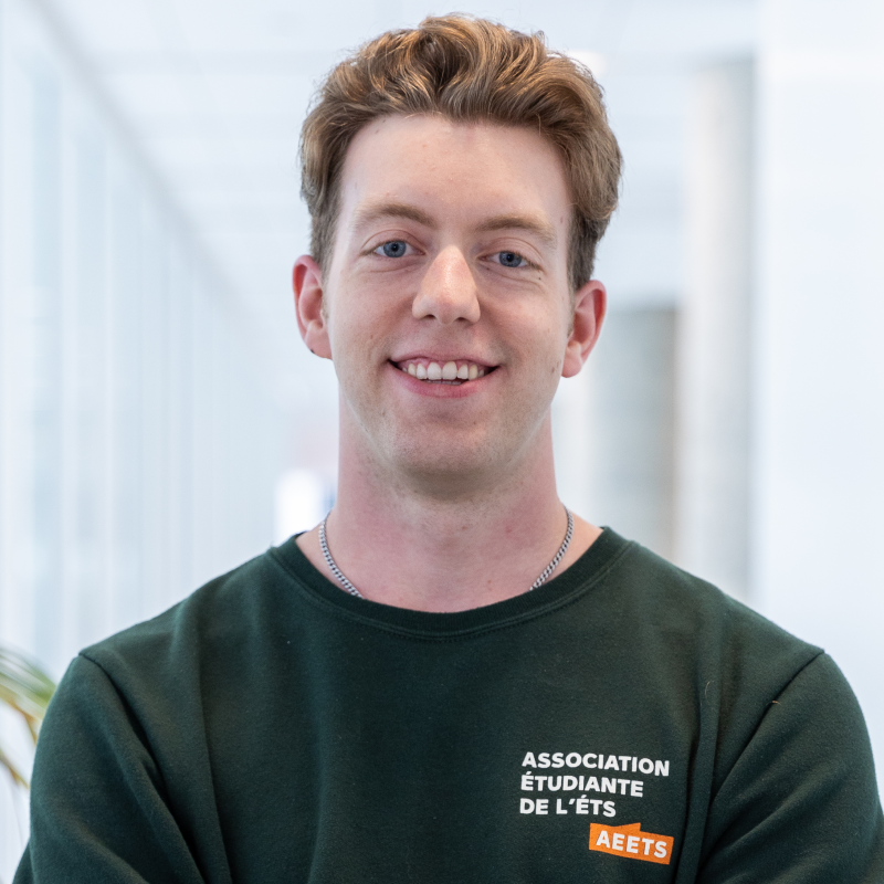 Un étudiant souriant portant un chandail de l'association étudiante de l'ÉTS, dans un environnement moderne et lumineux.