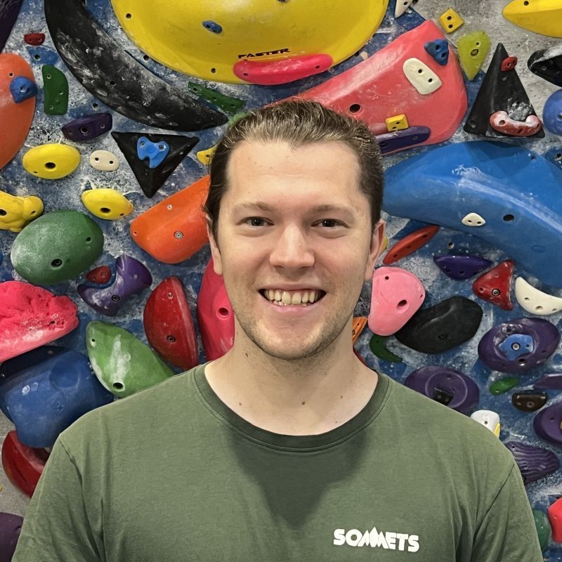 Un étudiant souriant en tenue décontractée se tient devant un mur d'escalade coloré, prêt pour l'aventure.